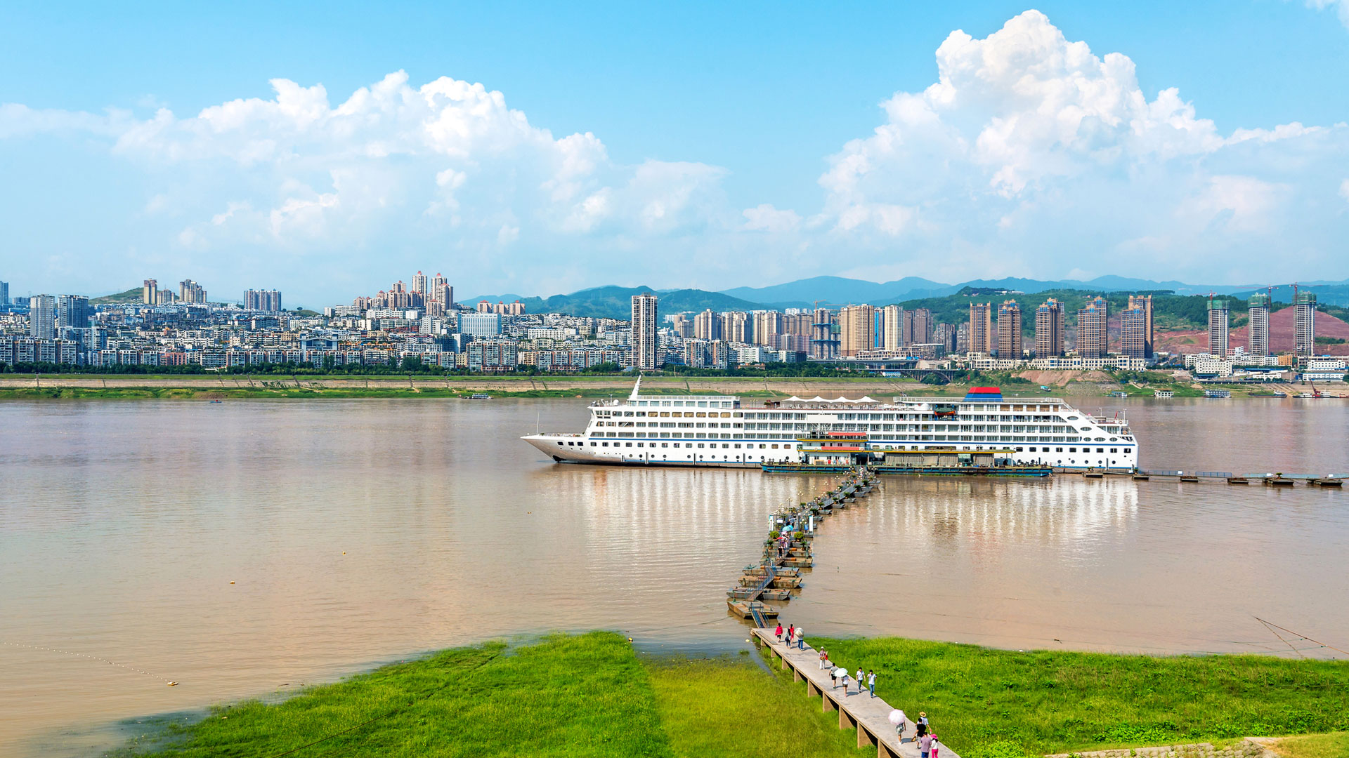 Cruise ship at a long pier walkway beneath a city skyline and summer clouds